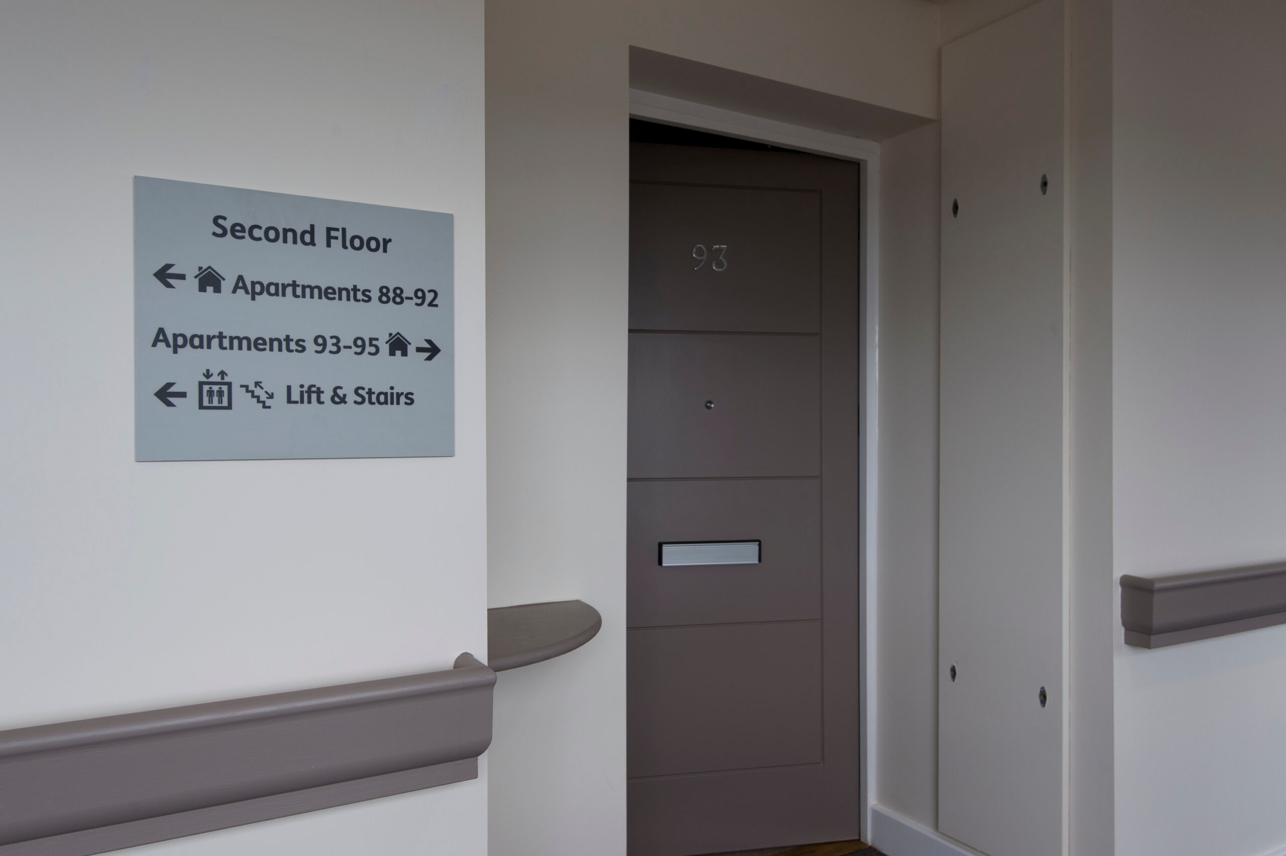 Interior corridor with a wayfinding sign on the wall reading “Second Floor,” with arrows directing to apartments 88–92, apartments 93–95, and lift and stairs. A numbered apartment door is visible beside the sign.