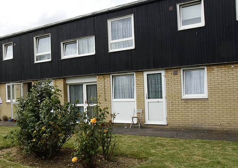 Exterior of a two‑storey residential building with light brick walls on the ground floor and black cladding on the upper floor. White‑framed windows and front doors face a small shared lawn with low shrubs and a paved path, with a chair placed outside one entrance.