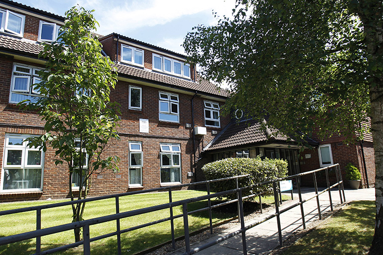 Exterior of a multi‑storey red‑brick residential building with white‑framed windows, viewed from a paved access ramp with metal handrails. A neatly maintained lawn, shrubs and mature trees surround the entrance, with a pitched‑roof canopy over the doorway and sunlight filtering through the foliage.