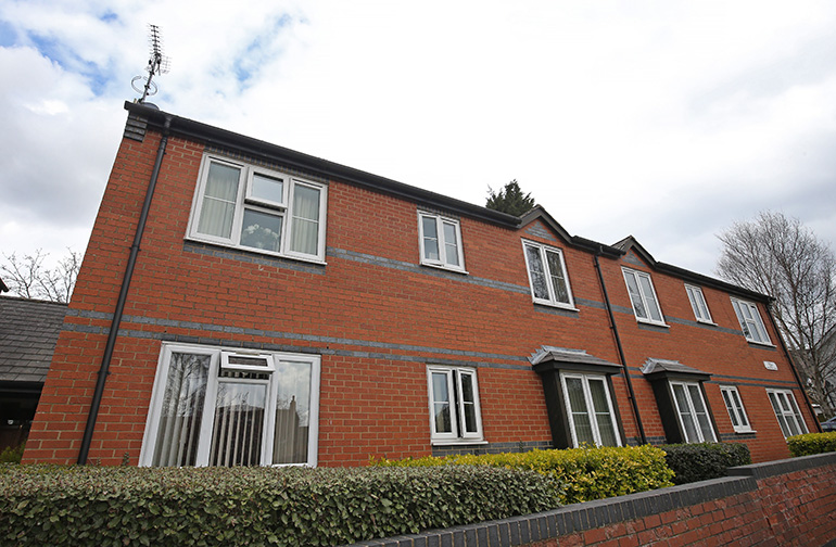 Exterior of a two‑storey red‑brick residential building with white‑framed windows and small entrance canopies. A low hedge and brick wall run along the front of the property beside a paved pavement, with neighbouring buildings and leafless trees visible under an overcast sky.