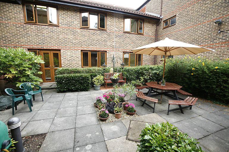 Communal courtyard outside a two‑storey brick residential building with white‑framed windows and wooden doors. A paved seating area includes benches, a picnic table with a large parasol, and potted plants arranged around the space, with hedges and shrubs creating a sheltered shared garden.