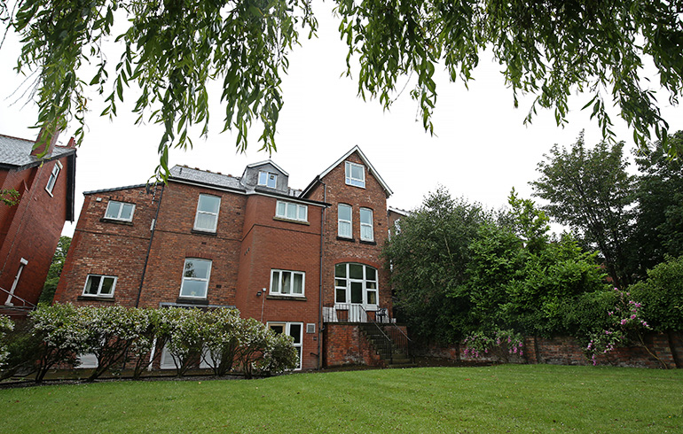 Exterior rear view of a multi‑storey red‑brick residential building with white‑framed windows, set behind a large grassy lawn. A central stepped entrance leads up to glass double doors, with hedges and flowering shrubs along the boundary and mature trees and greenery surrounding the garden area under an overcast sky.