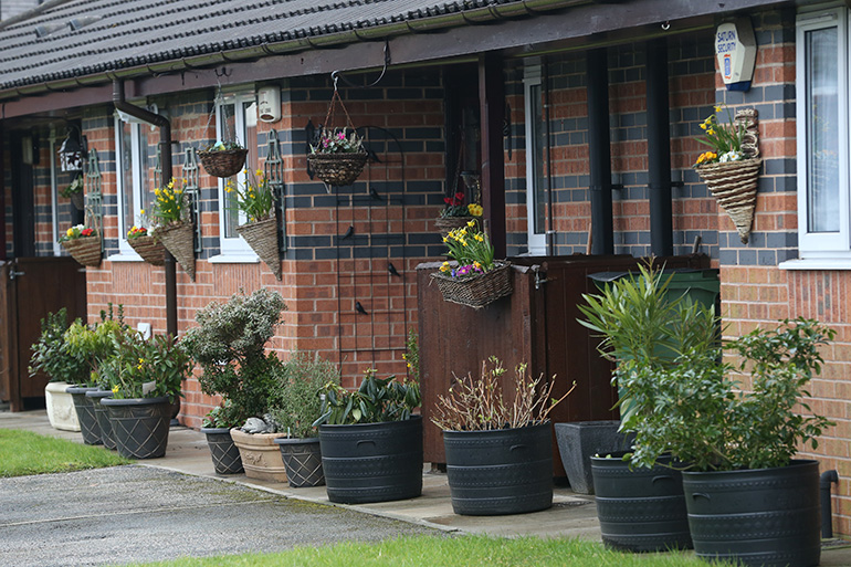 Close‑up of the exterior of a single‑storey red‑brick residential building, showing front doors and white‑framed windows. Hanging baskets and large potted plants filled with flowers and shrubs line the entrance areas, with a paved path and small lawn in the foreground.