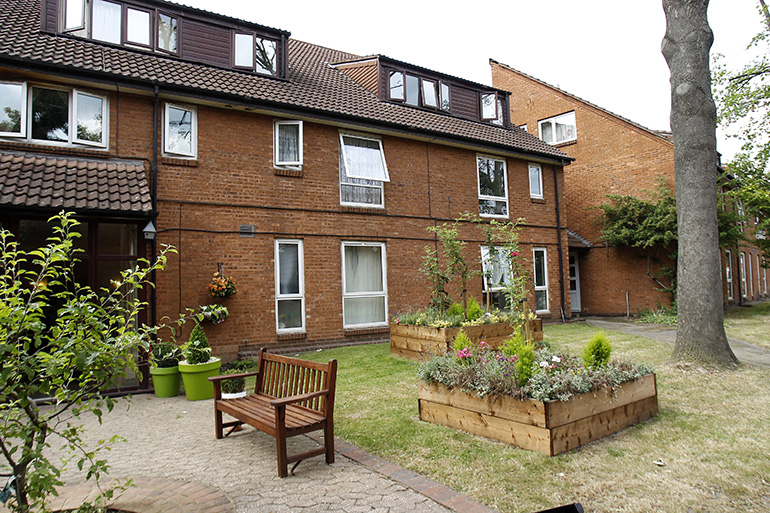 Communal garden courtyard in front of a multi‑storey red‑brick residential building with white‑framed windows. A wooden bench sits on a paved path alongside raised wooden planters filled with greenery, with lawn areas, potted plants and a mature tree creating a shared outdoor space beside the building under bright daylight.