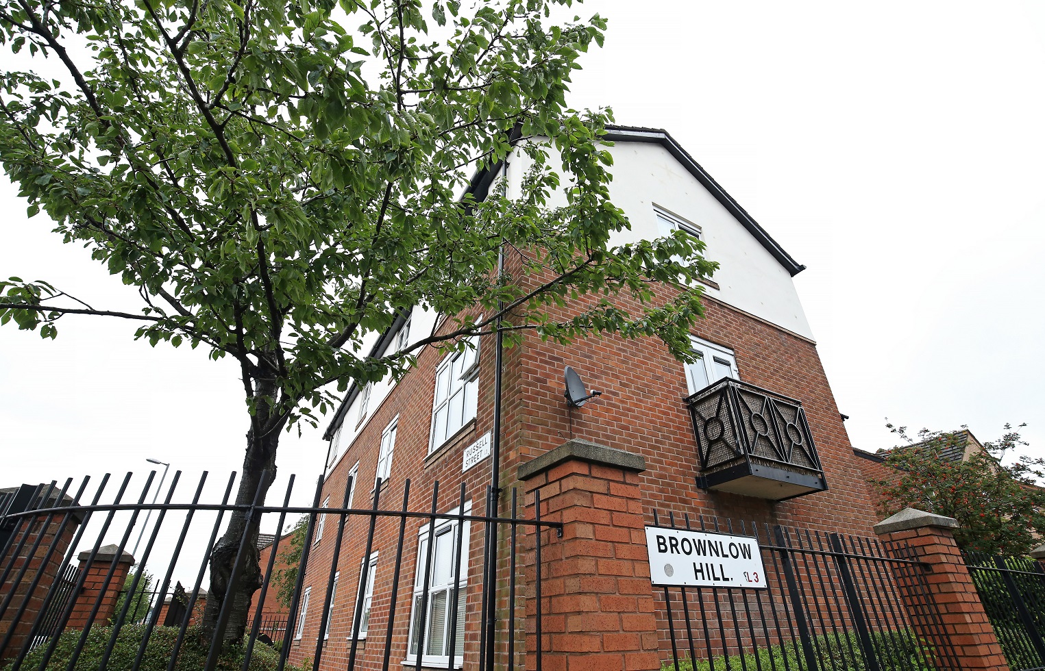Exterior of a multi‑storey red‑brick residential building with white‑framed windows, viewed from the pavement behind a low brick wall and dense green hedging. A tree with red berries stands in front of the building, with a metal fence along the boundary and neighbouring buildings visible under an overcast sky.