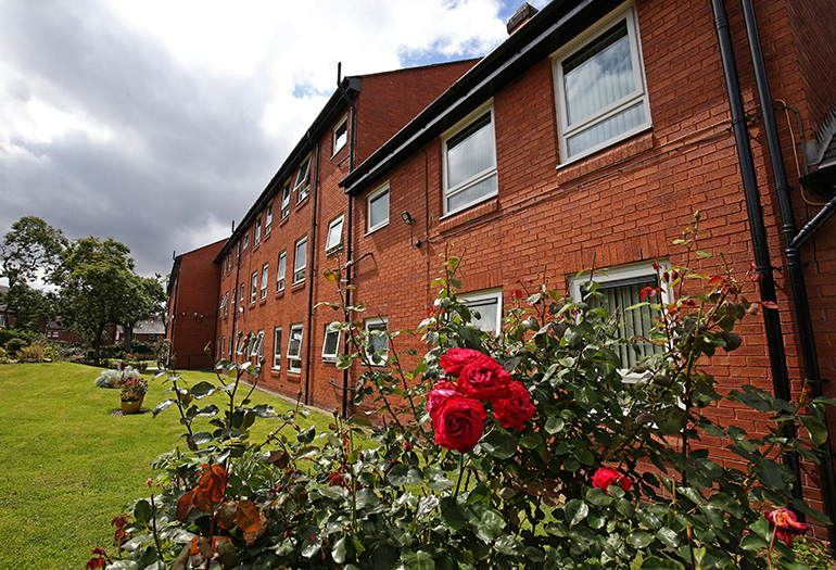 Exterior of a multi‑storey red‑brick residential building viewed across a landscaped garden. Red rose bushes are in bloom in the foreground, with a wide grass lawn, potted plants and white‑framed windows along the building under a cloudy sky.