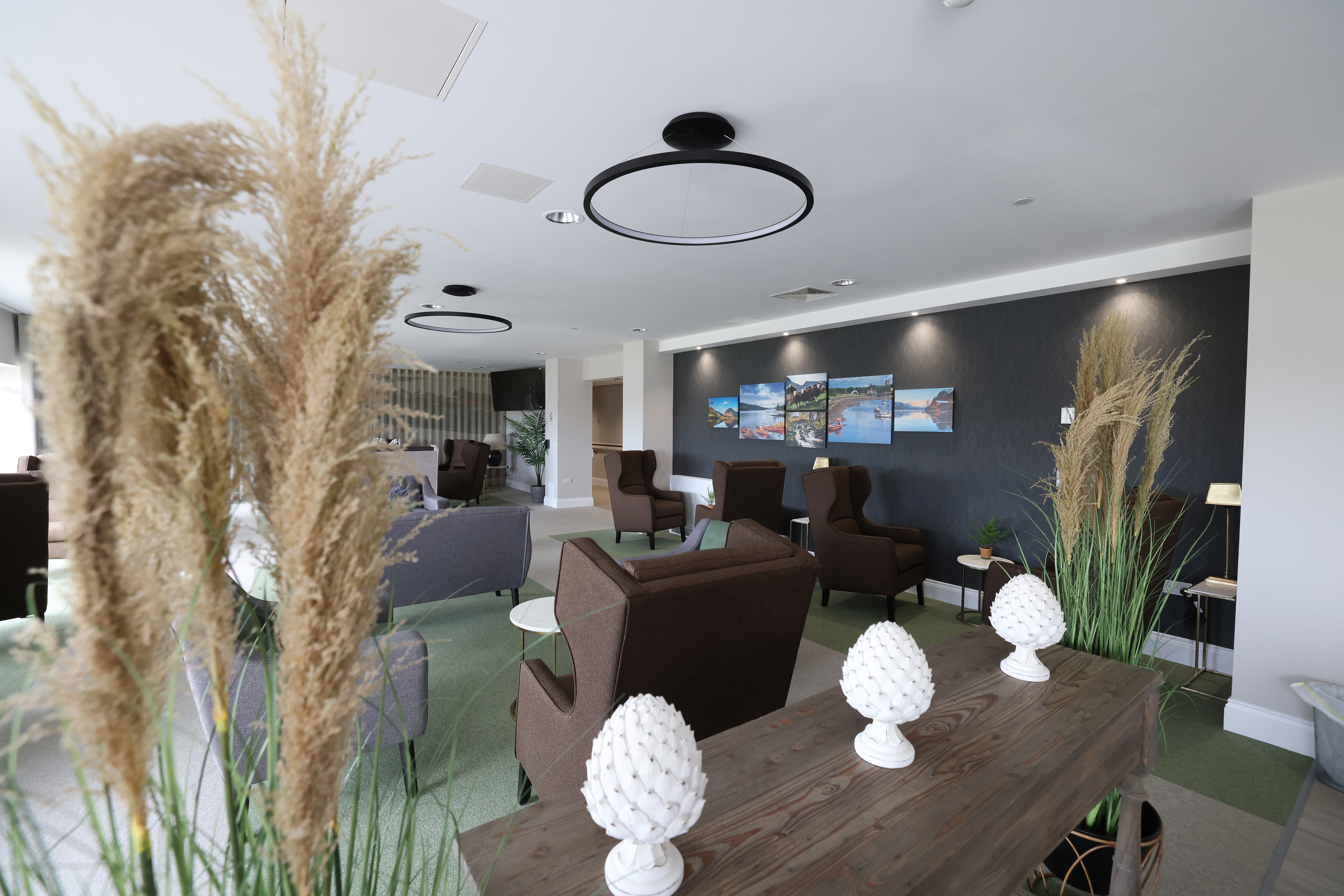 Bright communal lounge with armchairs arranged around small tables, decorative plants and modern ceiling lights. Framed landscape photographs hang on a dark feature wall, with carpeted flooring and soft lighting throughout.