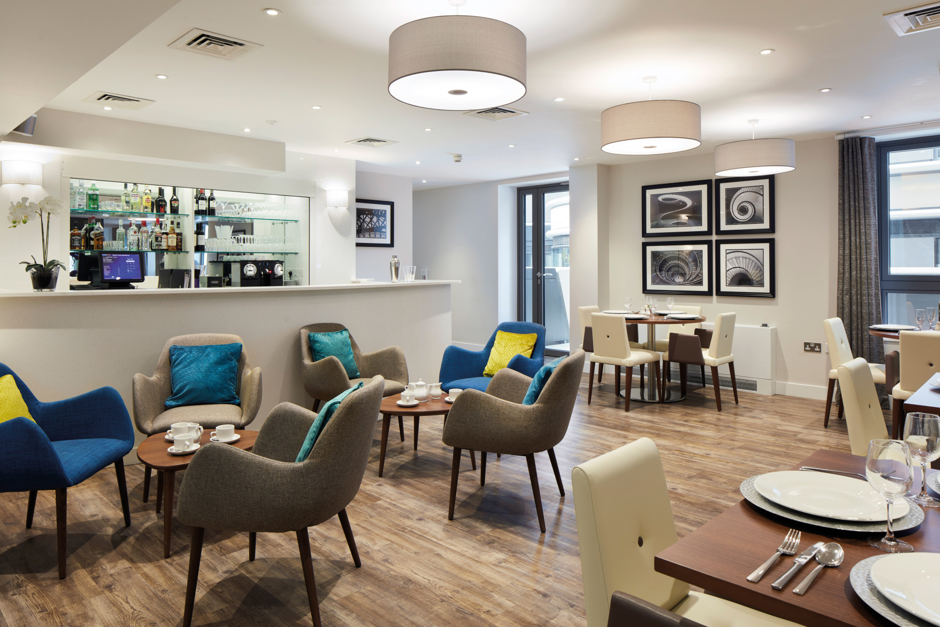 Communal dining and lounge area with round tables, upholstered chairs and wooden flooring. A bar counter with glass shelving and drinks sits at the back, with artwork on the walls and large ceiling lights creating a bright, modern interior.