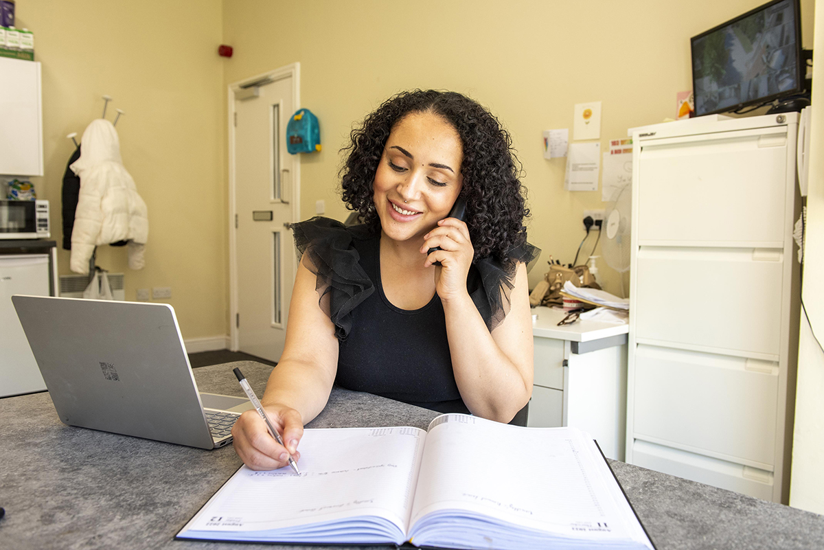 A woman sitting at a desk talking on the phone while writing in an open planner, with a laptop beside her.