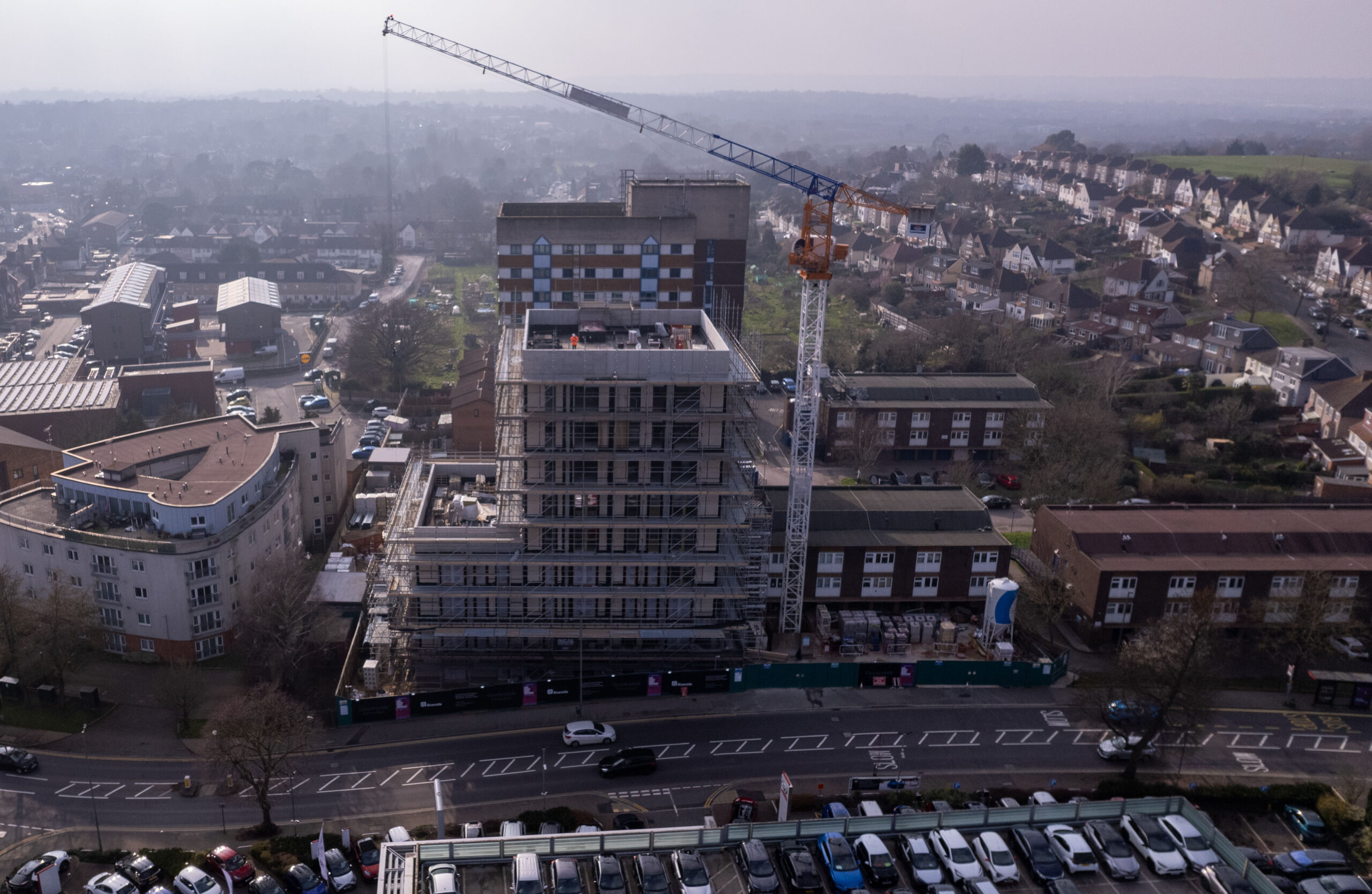 Aerial view of a multi‑storey building under construction with scaffolding and a crane beside it. The building is surrounded by residential streets, parked cars and neighbouring housing, with a townscape visible in the background.