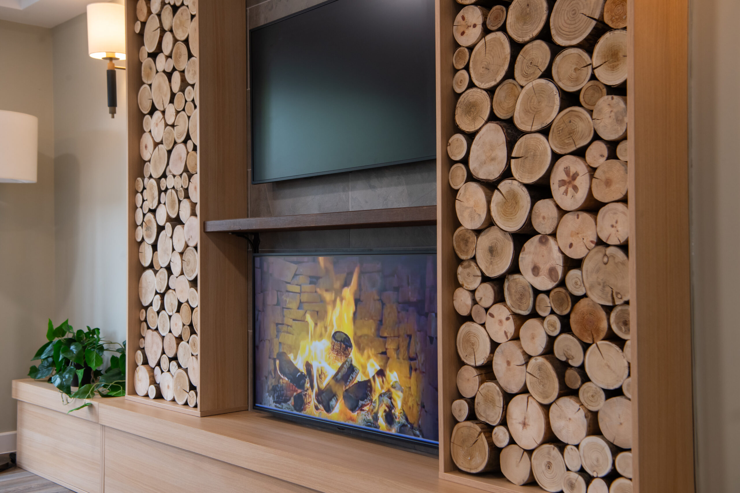 Modern interior feature wall with a realistic image of a built‑in fireplace showing a lit flame, framed by decorative stacked wood logs. A wall‑mounted television is positioned above the fireplace, with a plant and soft lighting to one side.