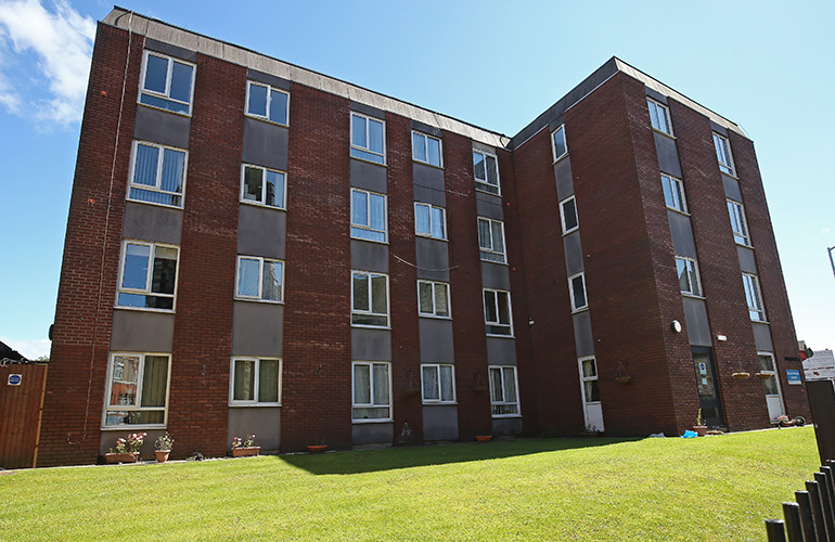 Exterior of a four‑storey red‑brick residential building with vertical grey panels between rows of white‑framed windows. A large, neatly maintained grass lawn stretches across the foreground, with potted plants along the base of the building and a bright blue sky overhead.