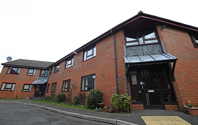 Exterior of a two‑storey red‑brick residential building with white‑framed windows and a main entrance beneath a metal canopy. The building sits along a quiet access road, with a gently sloping pavement, planted shrubs and pots near the entrance, and additional connected buildings visible in the background under an overcast sky.