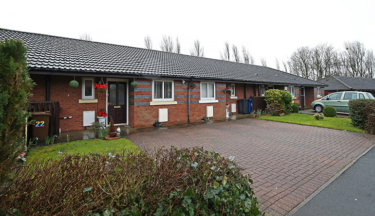 Exterior of a single‑storey red‑brick residential building with white‑framed windows and a dark tiled roof, arranged along a paved access road. Small front gardens with shrubs and potted plants sit outside each entrance, with a parked car to the right and mature trees in the background under an overcast sky.