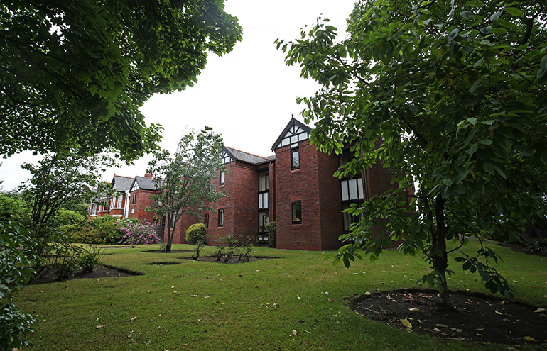 Exterior of a red‑brick residential building set within landscaped gardens, viewed through leafy trees in the foreground. The building has dark wood-framed windows and decorative gable detailing, with a well‑kept lawn, shrubs and flowering bushes surrounding the shared green space under an overcast sky.