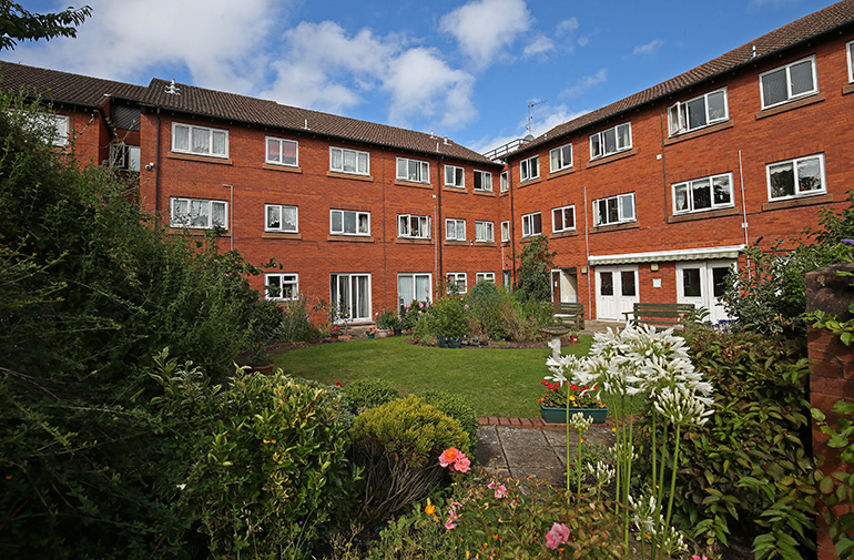 Communal garden courtyard surrounded by a multi‑storey red‑brick residential building with white‑framed windows. A central lawn and landscaped flower beds with shrubs and flowering plants fill the space, with a paved path leading toward the building entrance and mature planting around the edges under a partly cloudy blue sky.