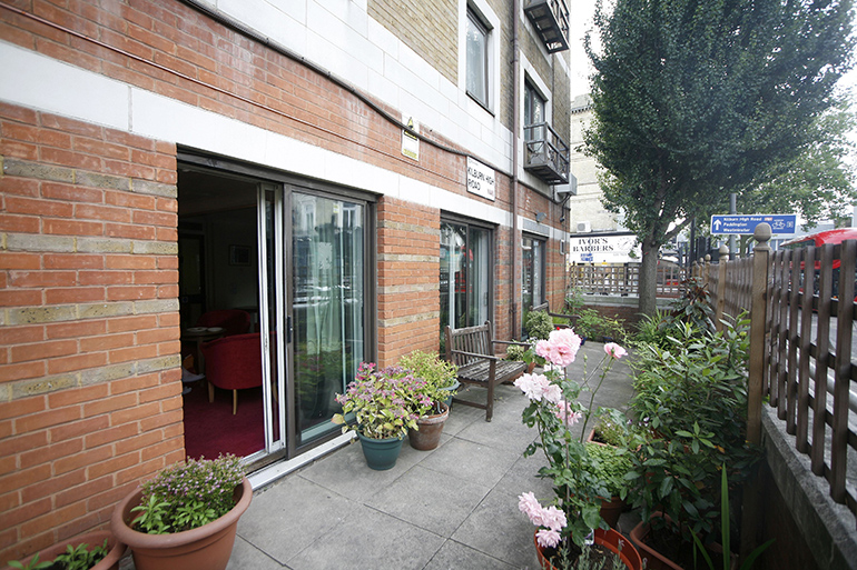 Exterior of a modern brick residential building with ground‑floor apartments opening onto a paved patio area. Glass sliding doors lead inside, with potted plants, pink flowers and a wooden bench arranged along the walkway. A fenced boundary with shrubs and a mature tree runs alongside the path, with neighbouring buildings and street signage visible in the background under overcast daylight.