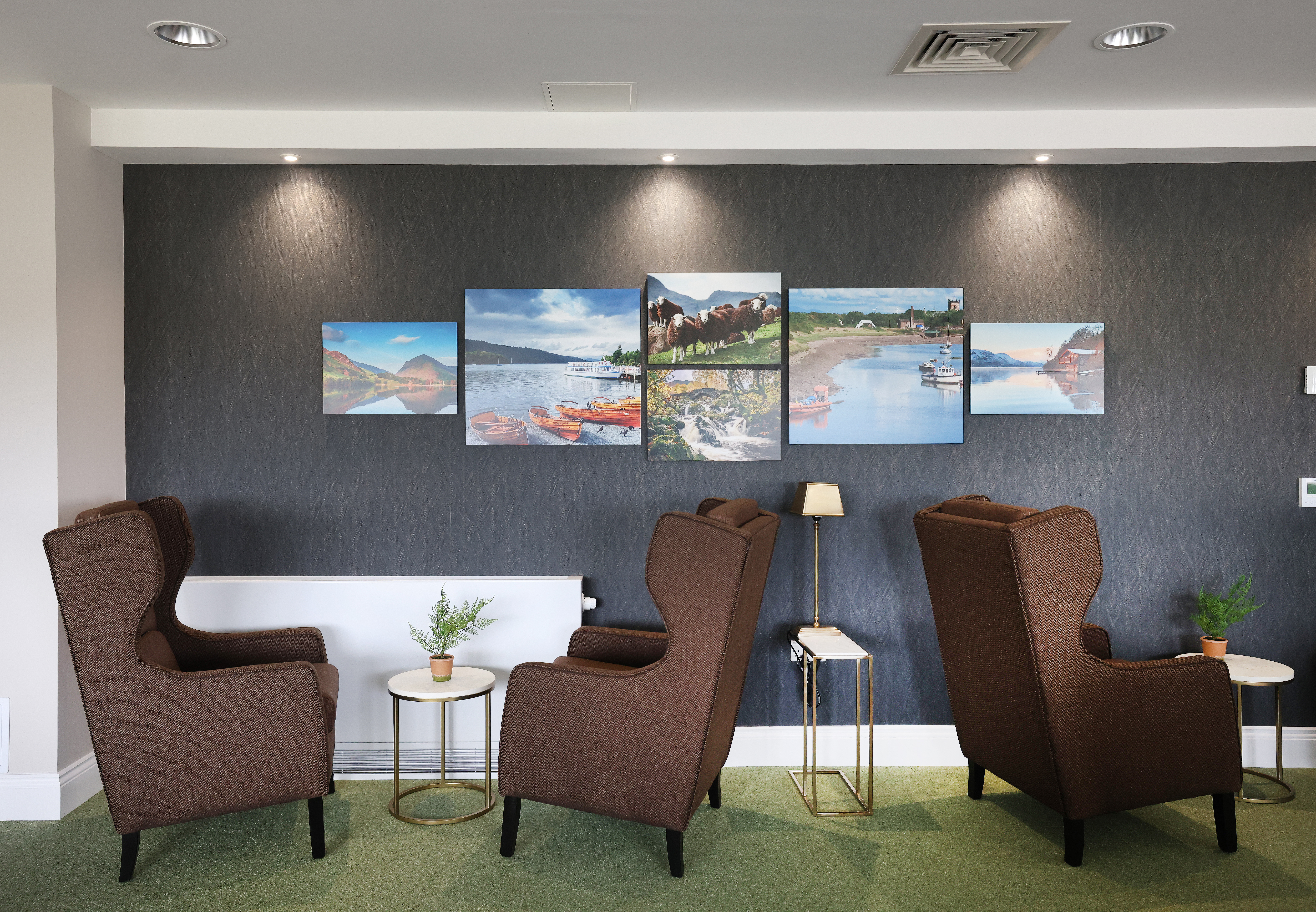 Row of high‑back armchairs positioned along a dark feature wall, with small side tables and lamps between them. Framed photographs of landscapes are mounted above the seating, creating a calm communal space.