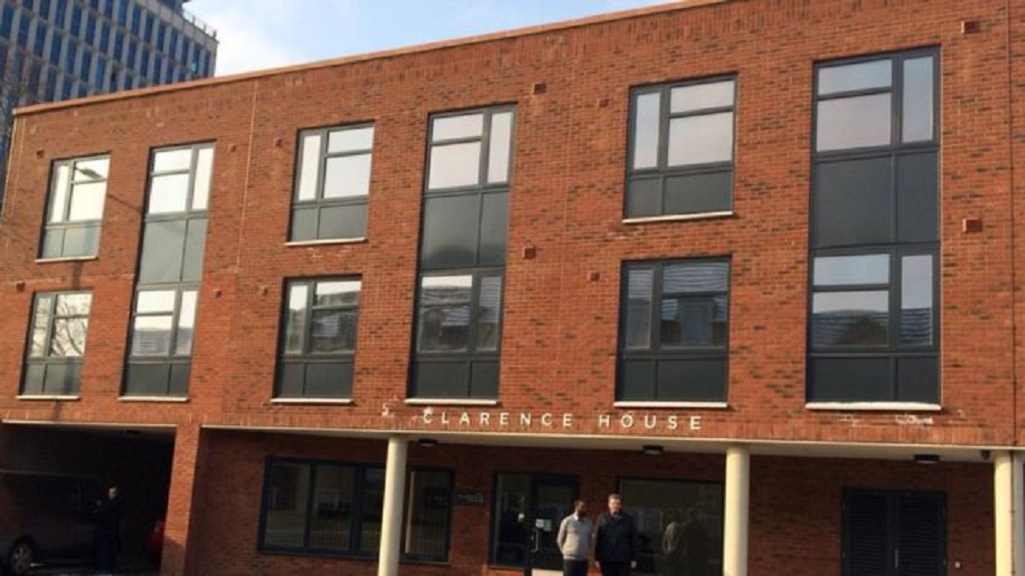 Exterior of a three‑storey red‑brick residential building with large black‑framed windows. A covered entrance at ground level is supported by columns and displays the name “Clarence House,” with paving in front and a taller office building visible in the background.