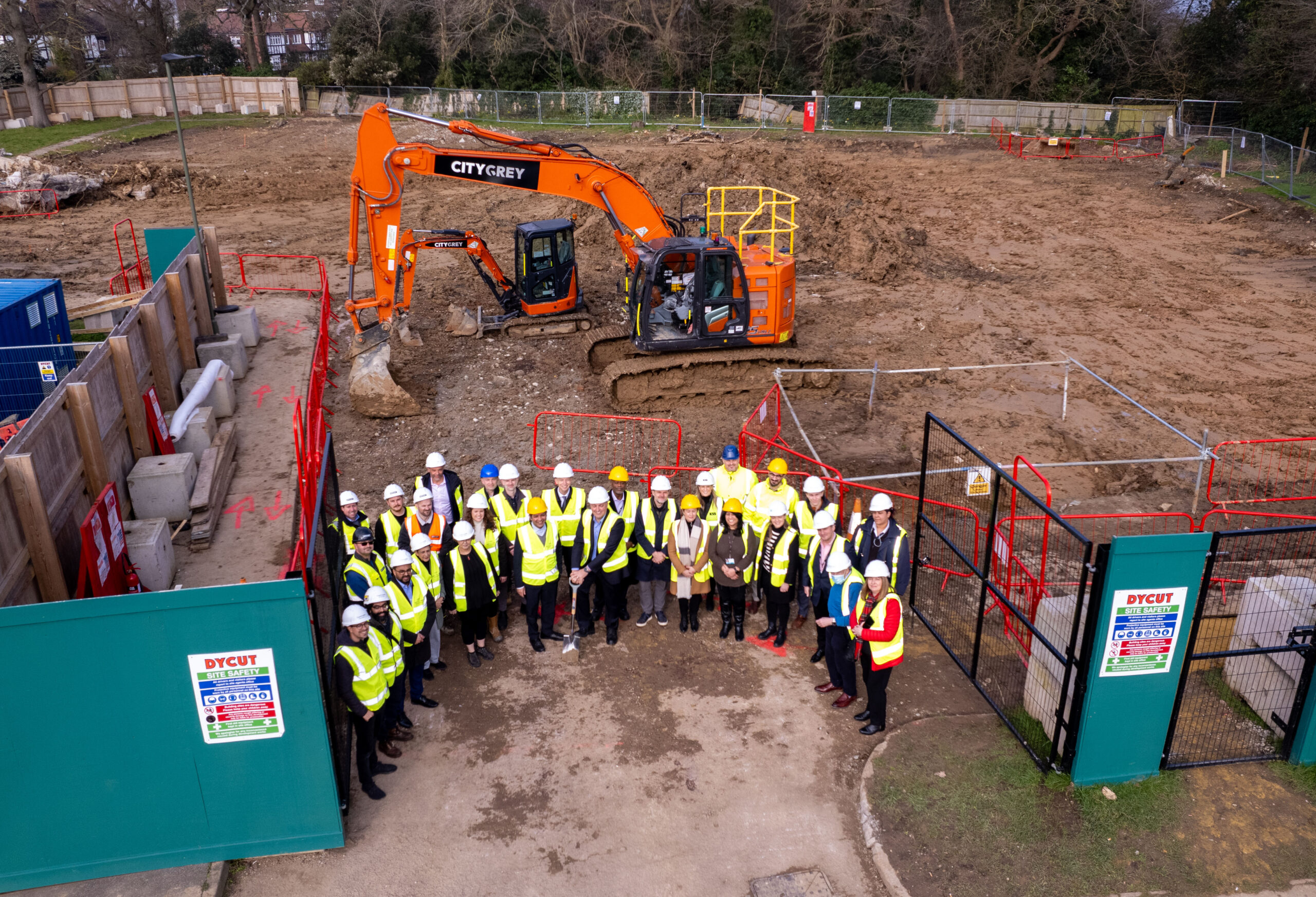 Group of people wearing high‑visibility vests and hard hats stand at a construction site during a ground‑breaking event. An excavator is positioned behind them on cleared ground, with temporary fencing and site signage visible around the perimeter.
