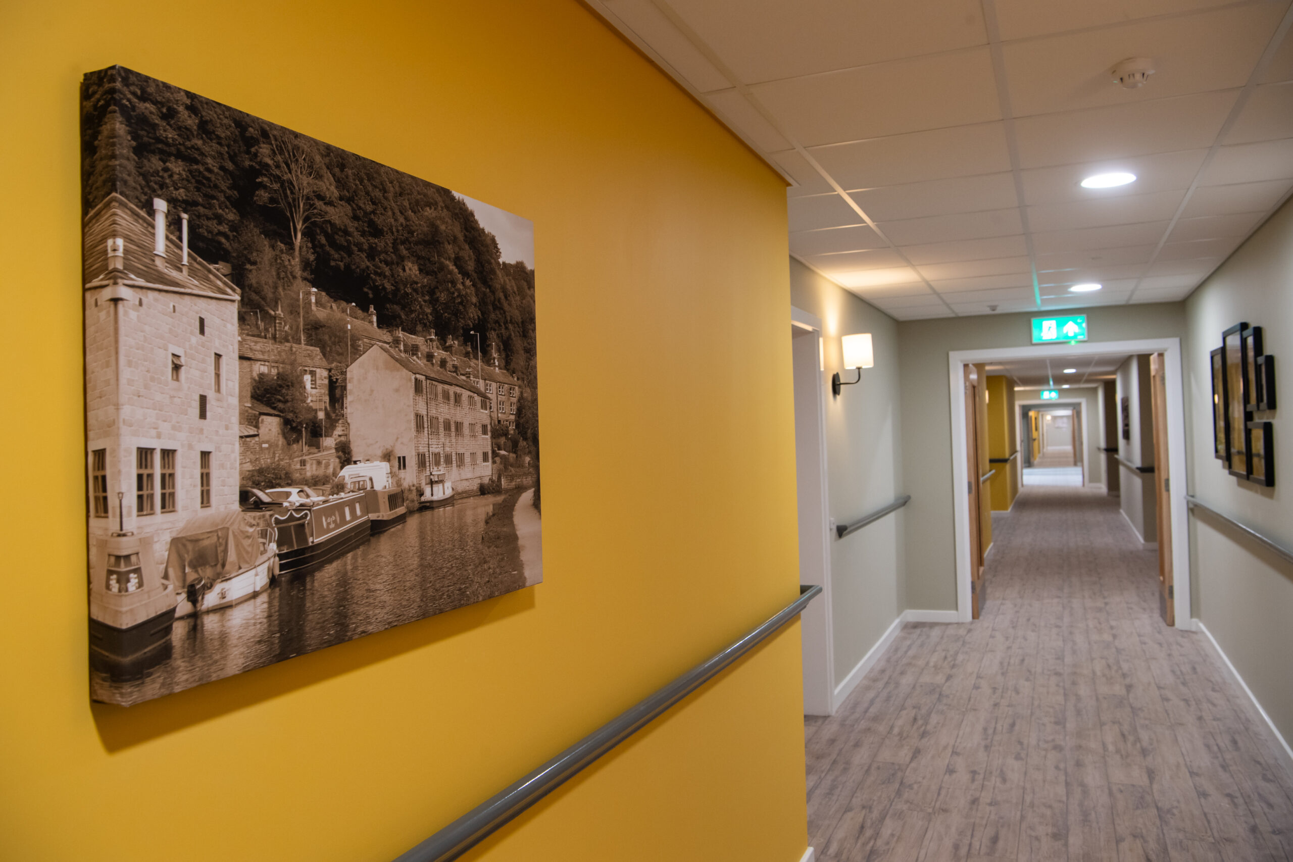 Interior hallway with a yellow feature wall displaying a framed black‑and‑white photograph of a canal and buildings. The corridor extends into the distance with handrails, ceiling lights and an illuminated exit sign.