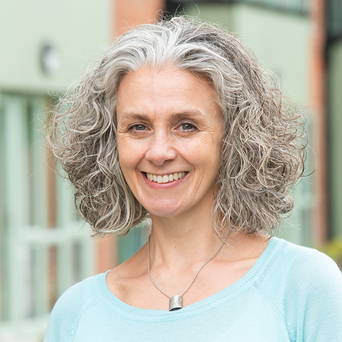 Portrait of a smiling woman with shoulder‑length curly grey hair wearing a light blue top. She stands outdoors in front of a modern building, with a softly blurred background.
