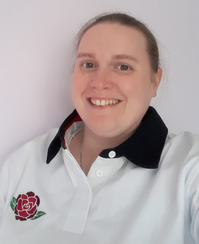 Head‑and‑shoulders portrait of a person facing the camera against a plain light‑coloured wall. They are wearing a white collared shirt with a dark collar detail and a rose emblem on the chest.