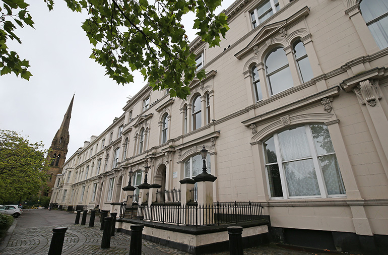 Exterior of a historic light‑stone residential building with ornate arched windows and decorative detailing along the façade. Black railings line the pavement in front, with trees overhead and a church spire visible further down the street.
