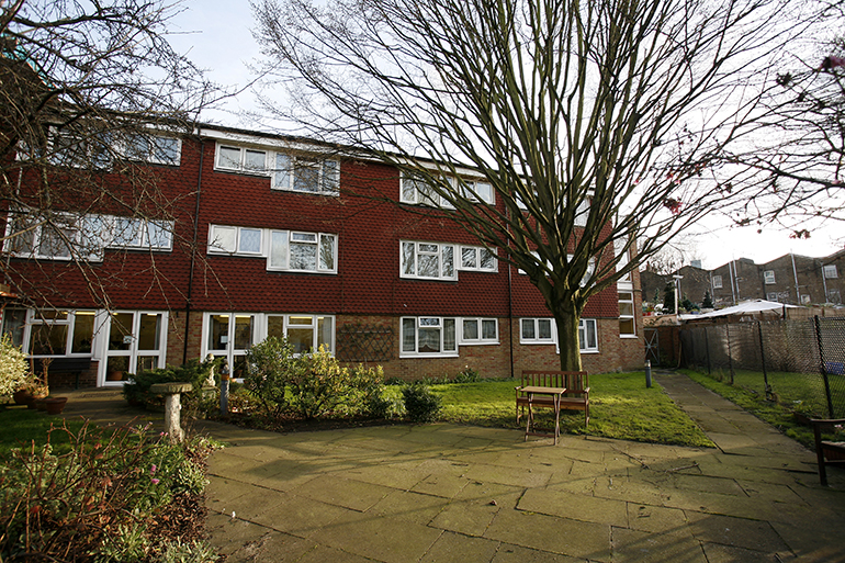Exterior of a red‑brick residential building with white‑framed windows, set around a paved communal courtyard. A wooden bench sits beside planted shrubs and a mature leafless tree, with lawns, pathways and neighbouring buildings visible beyond the shared outdoor space.