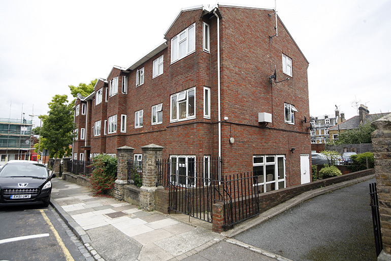 Exterior of a multi‑storey red‑brick residential building on a corner plot, with white‑framed windows and a pitched roof. A low brick wall and black metal railings border the frontage beside a pavement, with a parked car on the street and neighbouring buildings visible under an overcast sky.