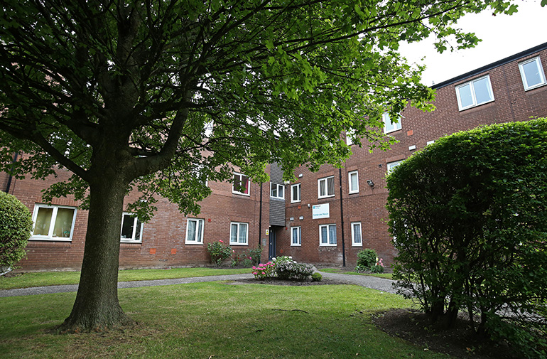 Exterior of a multi‑storey red‑brick residential building set around a landscaped communal garden. A large mature tree stands in the foreground beside a neatly kept lawn, with paved paths, shrubs and flower beds around the garden and white‑framed windows overlooking the shared outdoor space under an overcast sky.