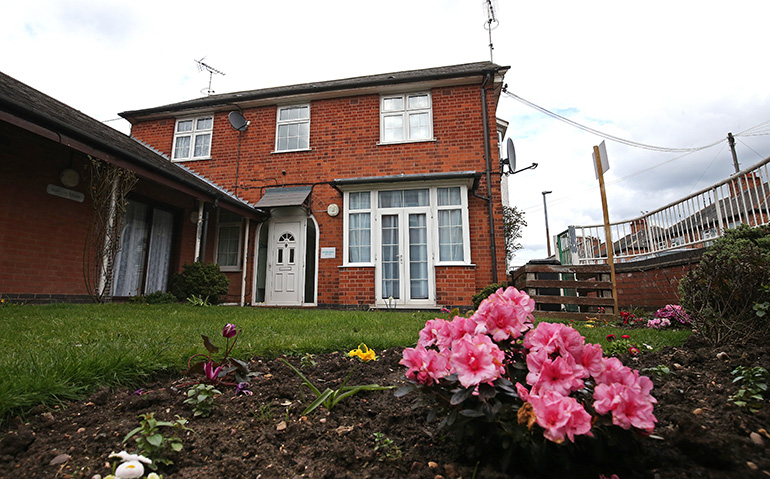 Exterior of a two‑storey red‑brick residential building with white‑framed windows and a central front door. A small front garden with a neatly kept lawn and a flower bed of pink and yellow flowers sits in the foreground, with steps and railings leading up to a raised walkway on the right and neighbouring buildings visible under a cloudy sky.