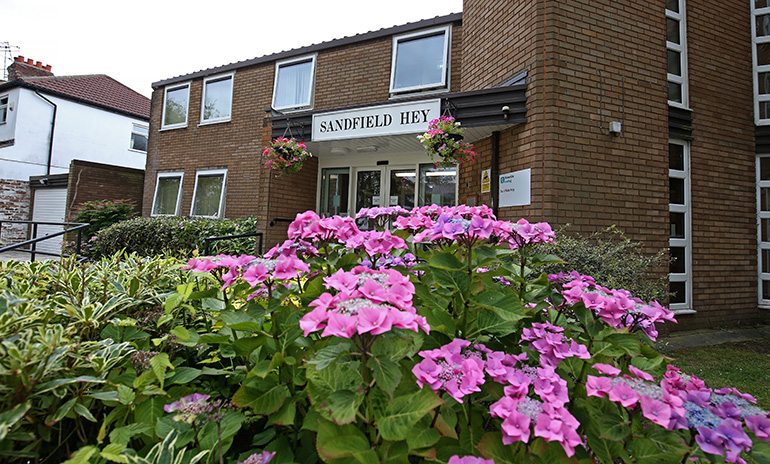 Exterior of a two‑storey light‑brick residential building with white‑framed windows and a central entrance. A sign above the doorway reads “Sandfield Hey,” with hanging baskets on either side. In the foreground, a landscaped garden features large pink flowers and green foliage, with shrubs and lawn around the entrance under overcast daylight.