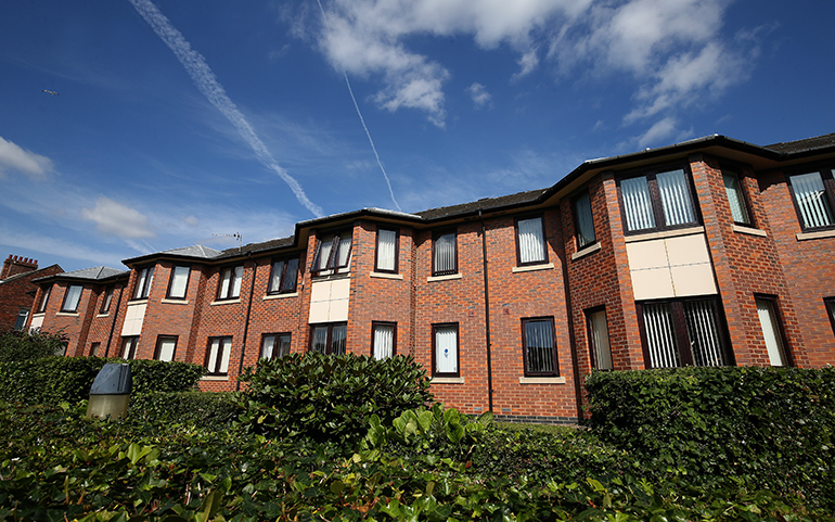 Exterior of a multi‑storey red‑brick residential building with cream‑coloured panels between rows of dark‑framed windows. A neatly trimmed hedge runs along the front of the building, with additional shrubs in the foreground and a bright blue sky with wispy clouds overhead.
