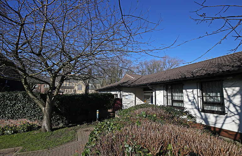 Exterior of a single‑storey light‑coloured residential building with a dark tiled roof, set within landscaped grounds. A paved path runs between trimmed hedges and shrubs, with a leafless tree and green lawn in the foreground and framed windows along the building under a clear blue sky.