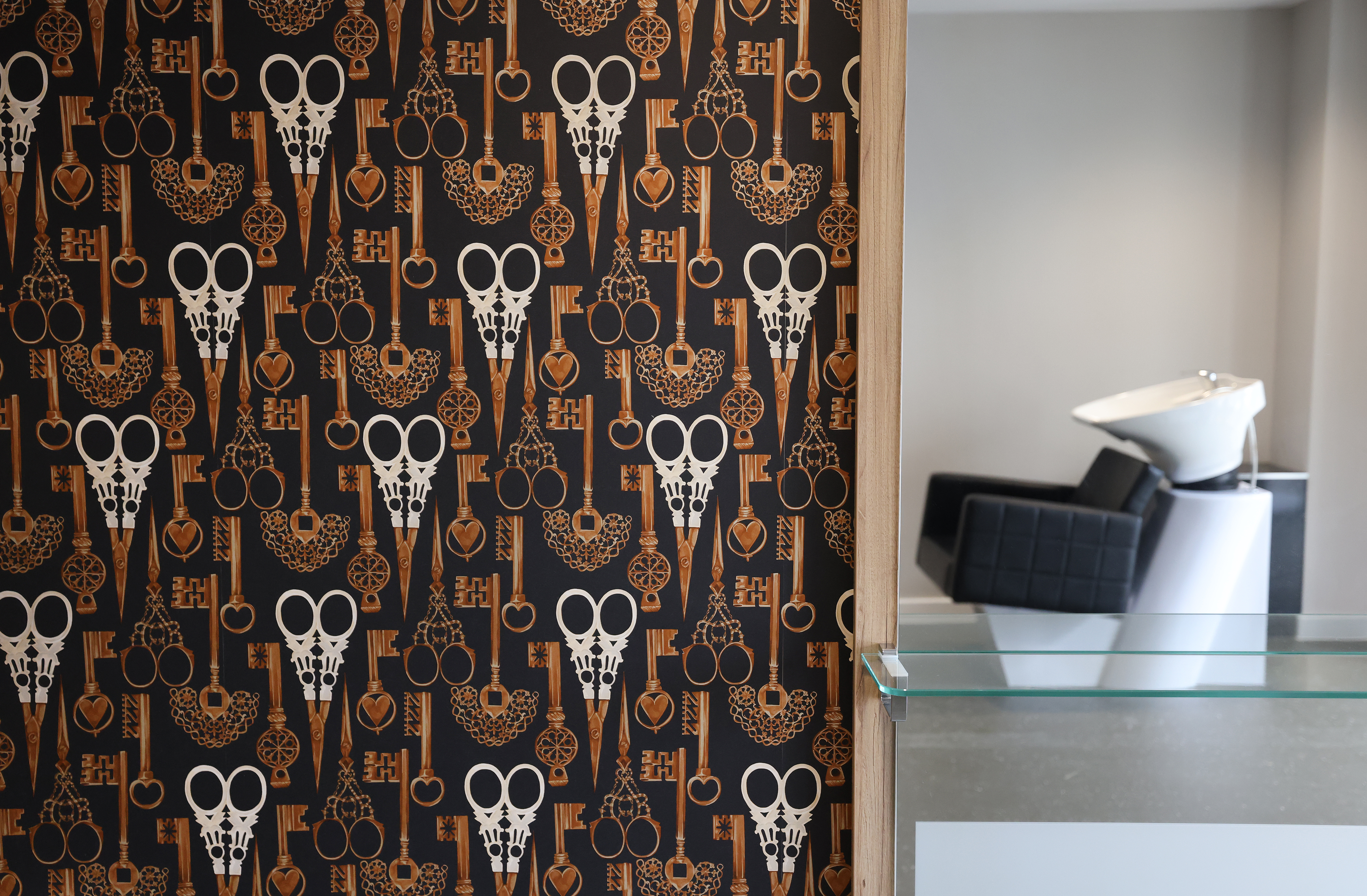 Close‑up of a decorative interior wall covered in patterned wallpaper featuring keys and scissors. A glass shelf and part of a modern white chair are visible to the right, highlighting interior design details.