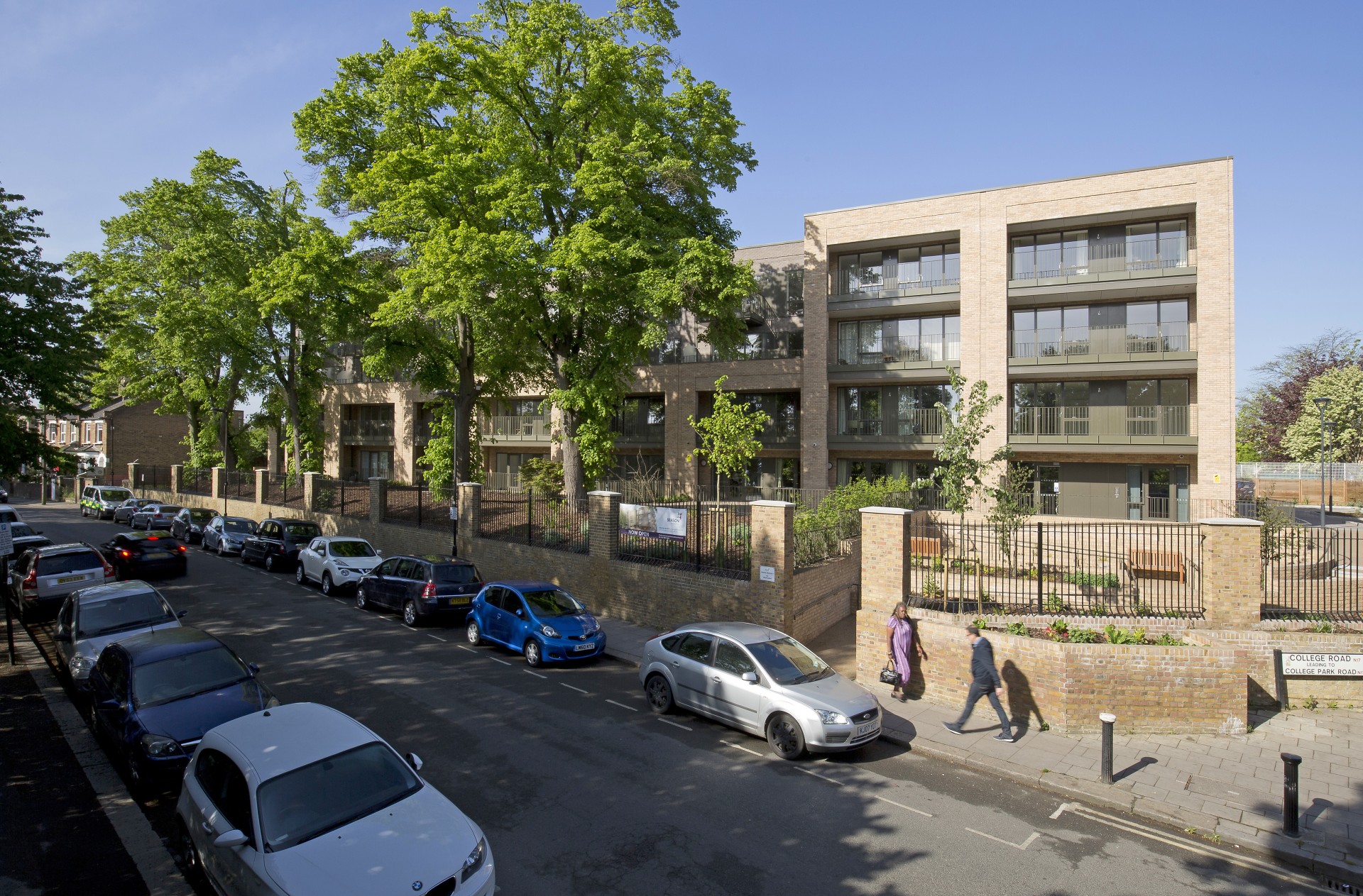 Street view of a modern multi‑storey residential development with light brick buildings and glass‑fronted balconies set behind a black metal fence. Mature green trees line the pavement, with parked cars along the road and two pedestrians walking beside the building under a clear blue sky.
