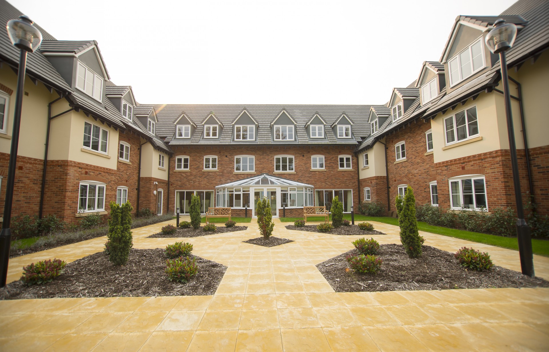 Enclosed courtyard surrounded by multi‑storey red‑brick residential buildings with dormer windows and cream panels. A paved central area features planted beds and young trees, with a glass‑roofed conservatory entrance and seating areas facing the landscaped space.