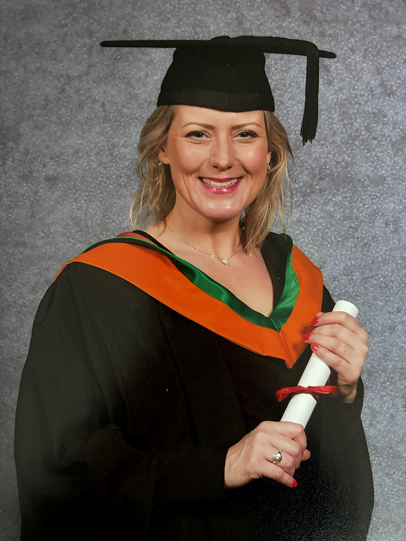 Women in Construction Week 2024_Millie Portrait of a smiling woman wearing graduation cap and gown, holding a rolled diploma certificate against a plain studio background.