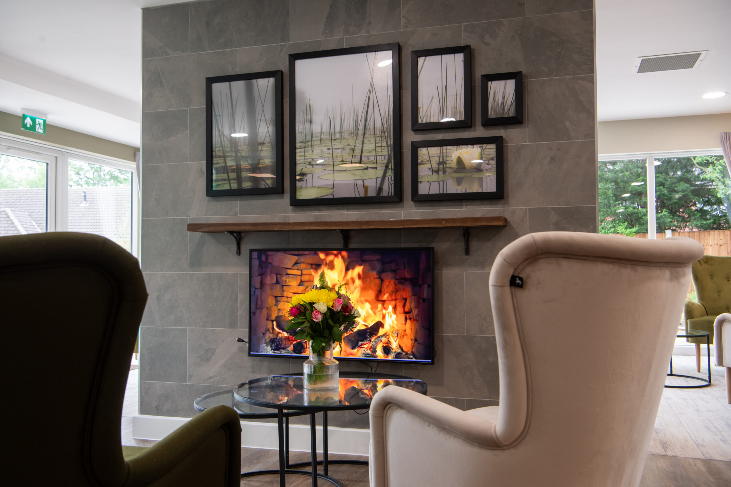 Comfortable communal lounge with armchairs arranged around a glass coffee table and a realistic image of a lit fireplace set into a tiled feature wall. Framed artwork hangs above the fireplace, and large windows bring natural light into the space.