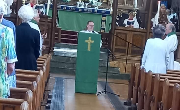 Interior of a church during a service, with a person standing at a lectern at the front, covered with a green cloth featuring a gold cross. Members of the congregation stand in wooden pews on either side of the aisle, with the altar and church furnishings visible in the background.