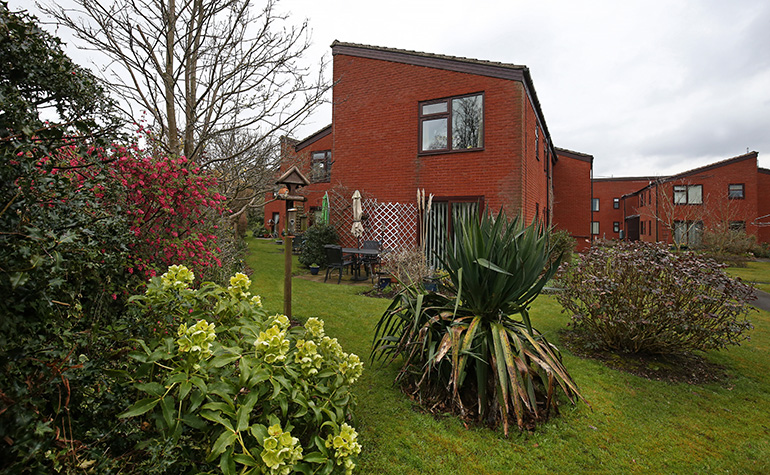 Landscaped communal garden beside red‑brick residential buildings, with a variety of shrubs and flowering plants in the foreground. A paved seating area with tables and chairs sits near trellis fencing, with a bird table on the lawn and additional residential blocks visible in the background under an overcast sky.