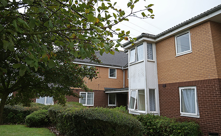 Exterior of a two‑storey light‑brick residential building with white‑framed windows and a pitched tiled roof. The building is set within landscaped grounds, with hedges, shrubs and a small lawn in the foreground, and a mature tree partially framing the view under overcast daylight.