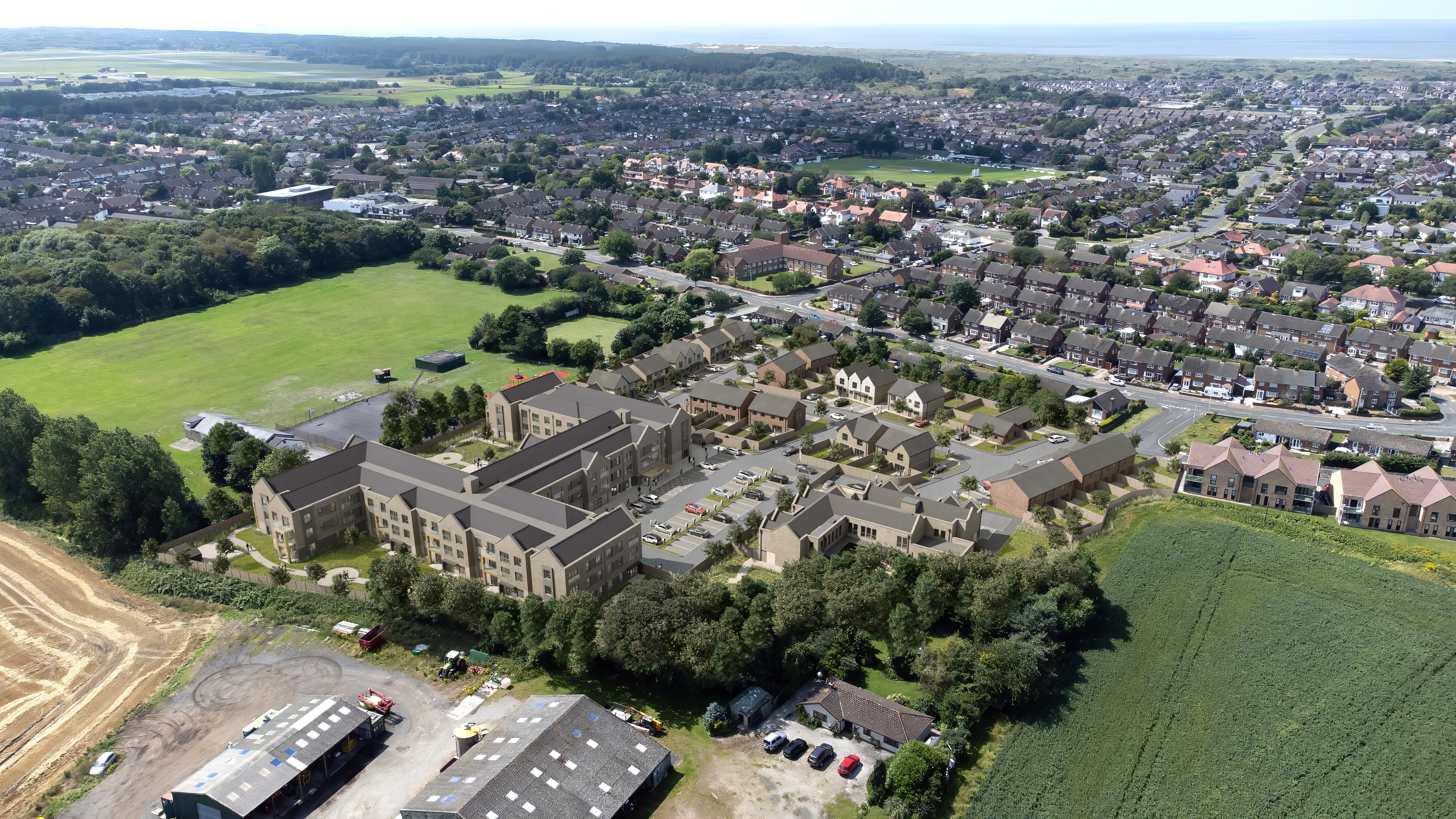 Aerial artist’s impression of a new residential development featuring clusters of modern homes and low‑rise apartment blocks arranged around shared streets and parking areas. The development sits beside an existing town with rows of houses, grassy fields, and woodland, with open countryside stretching toward the coastline in the distance.