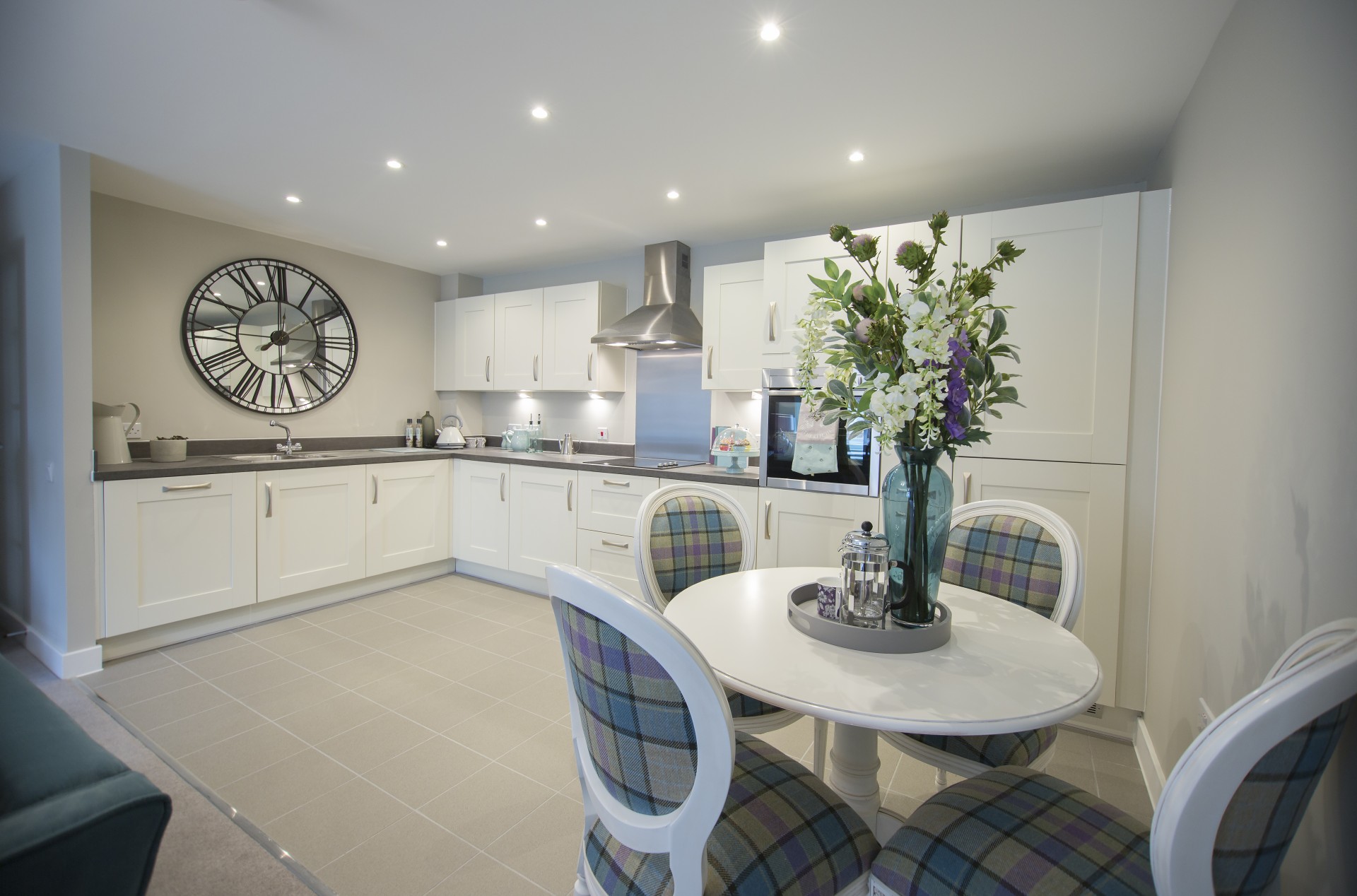 Modern open‑plan kitchen with white cabinets, grey worktops and integrated appliances. A round dining table with upholstered chairs sits in the foreground, decorated with a vase of flowers, under bright ceiling lighting.