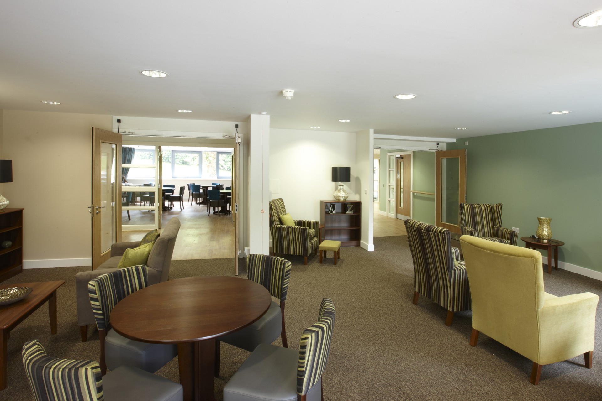 Communal lounge area with upholstered armchairs and sofas arranged around small tables on carpeted flooring. Double doors open into a dining room with tables and chairs, while a corridor extends to the right. Neutral décor, soft lighting and decorative lamps create a calm shared space.