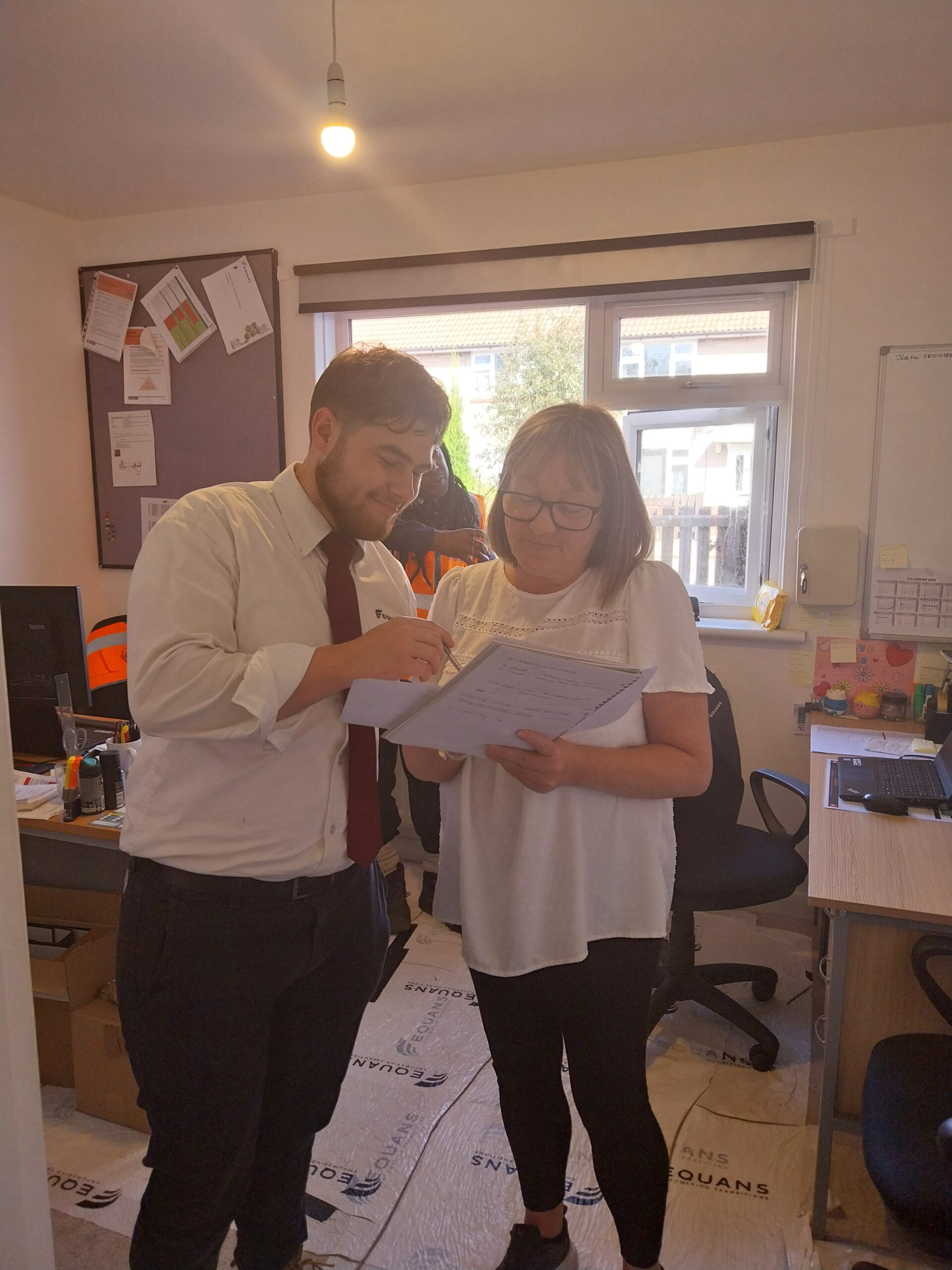 Two colleagues stand in an office looking at paperwork together, with one person pointing at the document while the other holds it. Desks, chairs and office noticeboards are visible in the background, with daylight coming through a window.