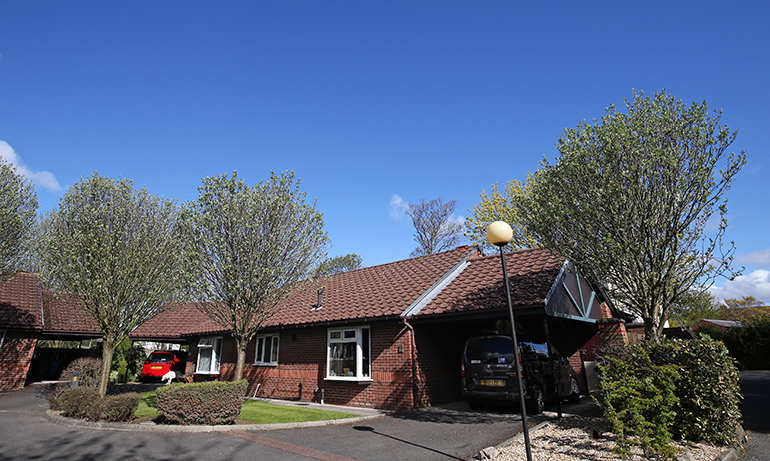 Exterior of a single‑storey red‑brick residential building with a pitched tiled roof, arranged around a small cul‑de‑sac. A carport extends from the building on the right, with parked vehicles beneath it. Neatly maintained lawns, trimmed shrubs and small trees surround the homes, with a clear blue sky overhead.