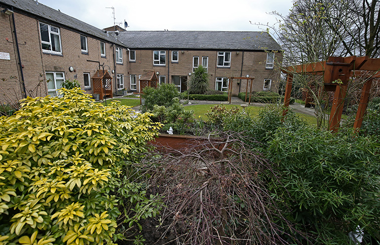 Landscaped communal garden in front of a two‑storey light‑brick residential building with white‑framed windows and doors. Shrubs and flowering plants fill the foreground, with raised planters, wooden pergola frames and a paved path leading through a well‑kept lawn under an overcast sky.