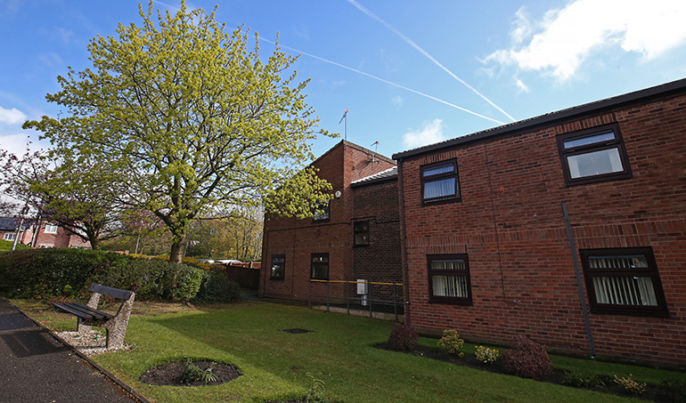 Landscaped communal garden beside a red‑brick residential building, with a neatly kept lawn, a paved path and a stone bench under a leafy tree. The building has wooden‑framed windows, with shrubs and small planted beds around the garden, set beneath a bright blue sky with light clouds.